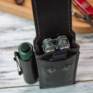 Puede incluir: Funda de cuero marr&oacute;n oscuro para multiherramientas con una linterna y una multiherramienta. La funda tiene un cierre de bot&oacute;n a presi&oacute;n y est&aacute; cosida con hilo marr&oacute;n m&aacute;s claro. Una linterna verde con clip negro est&aacute; a la izquierda.