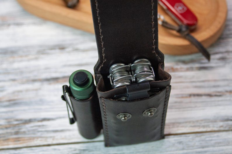 Puede incluir: Funda de cuero marr&oacute;n oscuro para multiherramientas con una linterna y una multiherramienta. La funda tiene un cierre de bot&oacute;n a presi&oacute;n y est&aacute; cosida con hilo marr&oacute;n m&aacute;s claro. Una linterna verde con clip negro est&aacute; a la izquierda.