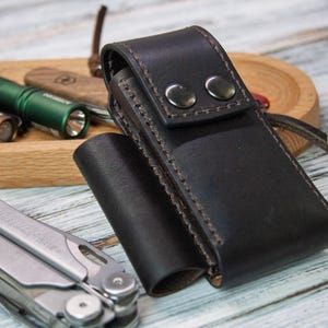 Puede incluir: Una funda de cuero negro para multiherramientas con dos cierres de bot&oacute;n a presi&oacute;n. Una multiherramienta plateada est&aacute; en primer plano, con una bandeja de madera que contiene linternas y una navaja en el fondo. Las linternas son marrones y verdes.