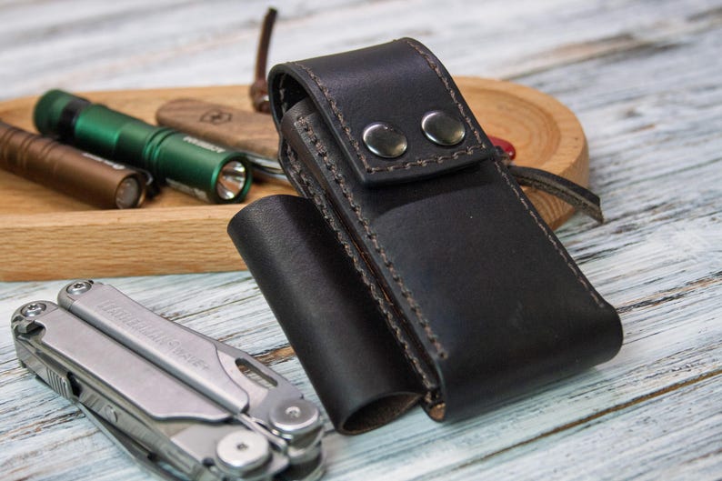 Puede incluir: Una funda de cuero negro para multiherramientas con dos cierres de bot&oacute;n a presi&oacute;n. Una multiherramienta plateada est&aacute; en primer plano, con una bandeja de madera que contiene linternas y una navaja en el fondo. Las linternas son marrones y verdes.