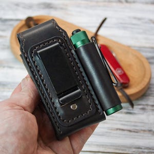 Puede incluir: Una funda de cintur&oacute;n de cuero negro con un soporte para linterna a juego. La funda tiene un clip de metal y est&aacute; cosida con hilo de color claro. Tambi&eacute;n se ven una linterna verde y negra y una navaja roja.