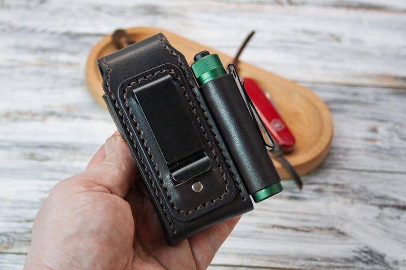 Puede incluir: Una funda de cintur&oacute;n de cuero negro con un soporte para linterna a juego. La funda tiene un clip de metal y est&aacute; cosida con hilo de color claro. Tambi&eacute;n se ven una linterna verde y negra y una navaja roja.