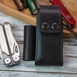 Puede incluir: Una funda de cuero negro para multiherramientas con dos cierres de bot&oacute;n a presi&oacute;n, una multiherramienta plateada y un objeto cil&iacute;ndrico negro. Una bandeja de madera contiene una navaja de bolsillo roja y varias linternas. La multiherramienta est&aacute; etiquetada como "LEATHERMAN WAVE +."