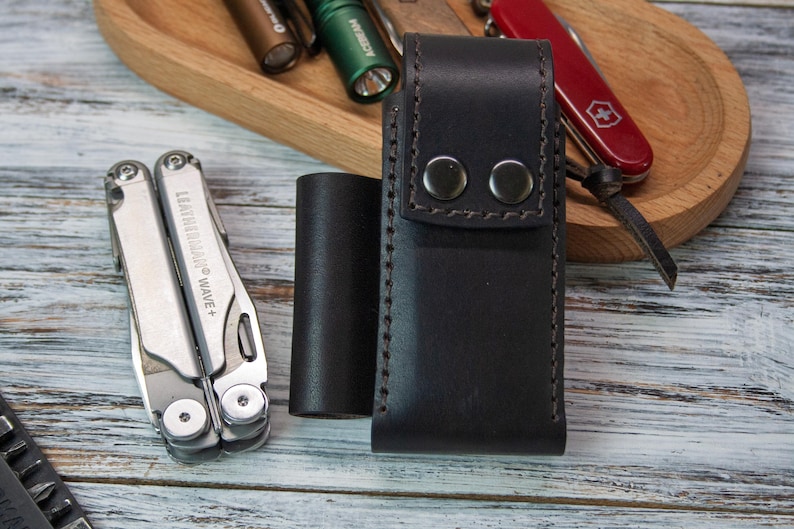 Puede incluir: Una funda de cuero negro para multiherramientas con dos cierres de bot&oacute;n a presi&oacute;n, una multiherramienta plateada y un objeto cil&iacute;ndrico negro. Una bandeja de madera contiene una navaja de bolsillo roja y varias linternas. La multiherramienta est&aacute; etiquetada como "LEATHERMAN WAVE +."