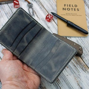 May include: A gray leather wallet with multiple card slots, featuring white stitching around the edges. The wallet is open, revealing the interior. A Field Notes notebook, pen, and dice are in the background.