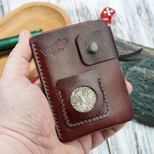 May include: A brown leather wallet with a silver coin detail. The wallet has a flap closure with a snap and is stitched around the edges. The "WayEdge Leather" logo is embossed on the front. A small, red Swiss Army knife and a green flashlight are in the background.