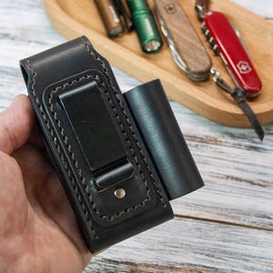 Puede incluir: Funda de linterna de cuero negro con un clip met&aacute;lico y una bolsa cil&iacute;ndrica separada. El soporte tiene costuras decorativas y un cierre a presi&oacute;n. Una bandeja de madera en el fondo contiene una variedad de herramientas peque&ntilde;as, incluido un cuchillo suizo rojo.