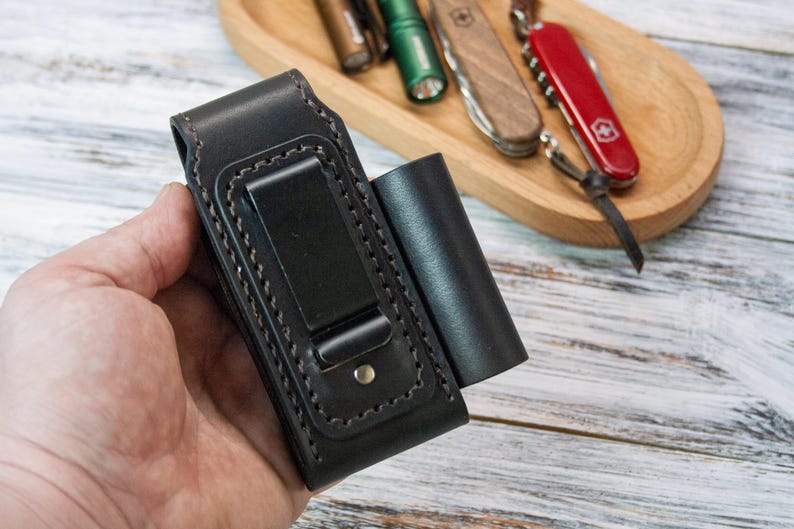 Puede incluir: Funda de linterna de cuero negro con un clip met&aacute;lico y una bolsa cil&iacute;ndrica separada. El soporte tiene costuras decorativas y un cierre a presi&oacute;n. Una bandeja de madera en el fondo contiene una variedad de herramientas peque&ntilde;as, incluido un cuchillo suizo rojo.