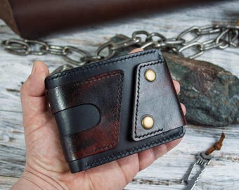 May include: A handmade leather wallet in black and burgundy. The wallet features a unique design with a flap secured by two brass-colored rivets. The interior and exterior are stitched with black thread. The wallet is held in a hand.