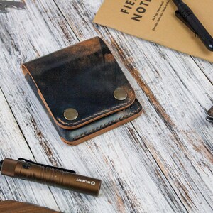 May include: A bi-fold wallet crafted from dark, distressed leather with a visible grain and two brass-coloured metal snap closures. The wallet features thick, dark stitching along the edges and a natural tan leather interior lining visible at the seams.