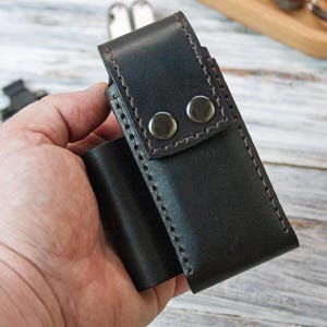 Puede incluir: Una funda de cuero marr&oacute;n oscuro con dos cierres de presi&oacute;n plateados. La funda est&aacute; cosida con un hilo en contraste y parece hecha a mano. La funda est&aacute; dise&ntilde;ada para guardar una herramienta peque&ntilde;a o un cuchillo. El art&iacute;culo se sostiene en la mano de una persona.