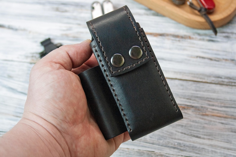 Puede incluir: Una funda de cuero marr&oacute;n oscuro con dos cierres de presi&oacute;n plateados. La funda est&aacute; cosida con un hilo en contraste y parece hecha a mano. La funda est&aacute; dise&ntilde;ada para guardar una herramienta peque&ntilde;a o un cuchillo. El art&iacute;culo se sostiene en la mano de una persona.