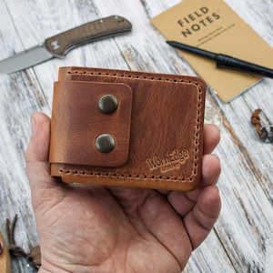 Peut inclure: Un portefeuille en cuir marron avec une fermeture à bouton-pression, avec le logo "WornEdge Leather". Le portefeuille est tenu dans une main, avec un couteau, un stylo et un bloc-notes en arrière-plan. Le portefeuille mesure environ 10 cm de large.