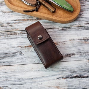 Puede incluir: Una funda de cuchillo de cuero marr&oacute;n oscuro con cierre de bot&oacute;n a presi&oacute;n. La funda es rectangular y parece estar cosida a mano. Otros art&iacute;culos, como un cuchillo y una linterna, son visibles en una bandeja de madera en el fondo.