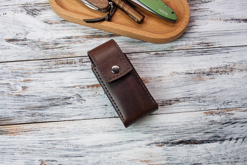 Puede incluir: Una funda de cuchillo de cuero marr&oacute;n oscuro con cierre de bot&oacute;n a presi&oacute;n. La funda es rectangular y parece estar cosida a mano. Otros art&iacute;culos, como un cuchillo y una linterna, son visibles en una bandeja de madera en el fondo.