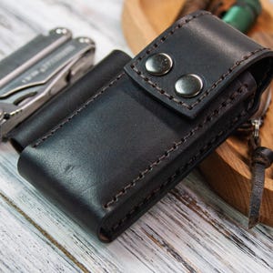 Puede incluir: Una funda de cuero negro para multiherramientas con dos cierres de bot&oacute;n plateados. Tambi&eacute;n se ven una multiherramienta plateada y una peque&ntilde;a navaja suiza roja. Los art&iacute;culos est&aacute;n sobre una superficie de madera clara.