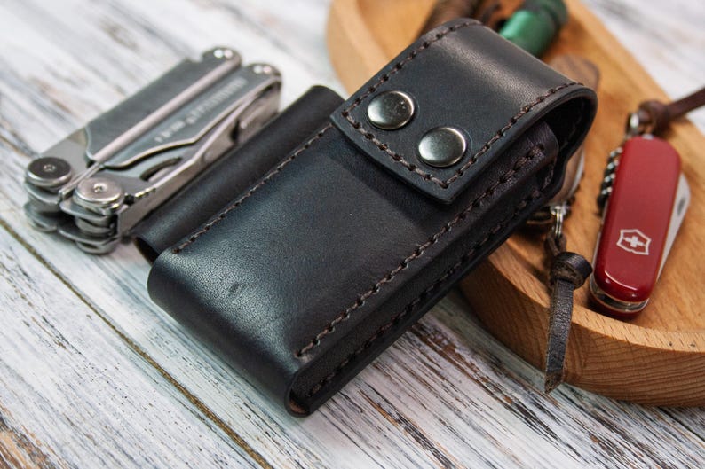 Puede incluir: Una funda de cuero negro para multiherramientas con dos cierres de bot&oacute;n plateados. Tambi&eacute;n se ven una multiherramienta plateada y una peque&ntilde;a navaja suiza roja. Los art&iacute;culos est&aacute;n sobre una superficie de madera clara.