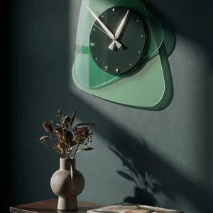 May include: A modern wall clock with a dark green face and silver hands, set against a layered, abstract green glass design. The clock is mounted on a dark teal wall. A vase with dried flowers and a book titled "Modernism" are on a nearby table.