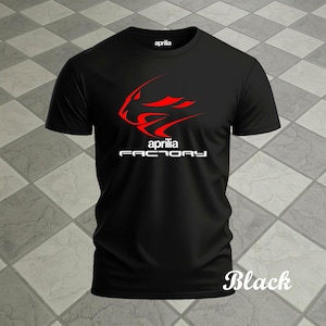 May include: Black t-shirt with a red and white graphic design. The design includes the word "aprilia" above a stylized image and the words "FACTORY" below. The word "Black" is written in a script font at the bottom right.