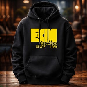 May include: Black hoodie featuring a yellow rectangular logo with the text "ECM RECORDS SINCE 1969". The sweatshirt has a front pocket and a drawstring hood. The background is blurred.