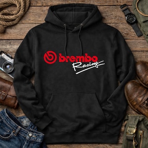 Pode incluir: Camisola com capuz preta com bolso canguru, com o logótipo "brembo Racing" em vermelho e branco. O design inclui um emblema vermelho estilizado acima do nome da marca. A camisola é exibida numa superfície de madeira com outros acessórios.