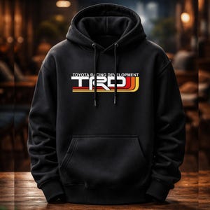 May include: Black hooded sweatshirt with a front pocket and drawstring. The front displays the text "TOYOTA RACING DEVELOPMENT" above the TRD logo in white, red, and yellow. The sweatshirt is made of a soft material.