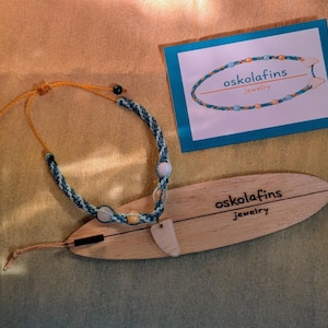 Könnte beinhalten: Eine handgefertigte Halskette mit einem blau-weißen geflochtenen Kordel, mit weißen und gelben Perlen und einem hellfarbigen Anhänger. Die Halskette wird auf einem surfbrettförmigen Holzdisplay mit dem Text "oskola fins jewelry" präsentiert. Eine Visitenkarte ist ebenfalls vorhanden.