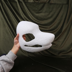 May include: A white, textured mask with large eye openings and a snout-like projection. The mask is held against a dark green background, showcasing its three-dimensional form and intricate design. The mask appears to be made of a porous material.