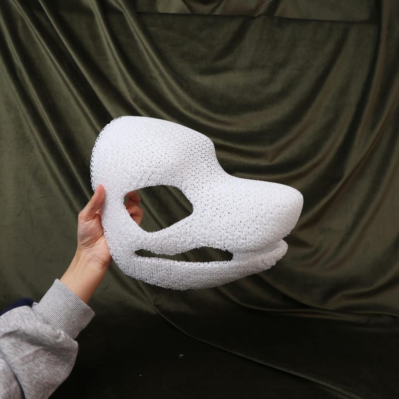 May include: A white, textured mask with large eye openings and a snout-like projection. The mask is held against a dark green background, showcasing its three-dimensional form and intricate design. The mask appears to be made of a porous material.