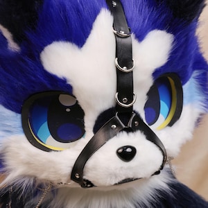 May include: A close-up of a furry, blue, white, and black animal mask with large, cartoon-style eyes. The mask has a black leather muzzle and a chain detail. The eyes are blue with yellow accents. The fur is a mix of blue, white, and black.