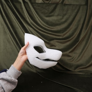 May include: A white, textured foam mask in the shape of an animal's face, featuring eye and mouth openings. The mask is held against a dark green, draped fabric background. The mask's design suggests a stylized animal or creature.