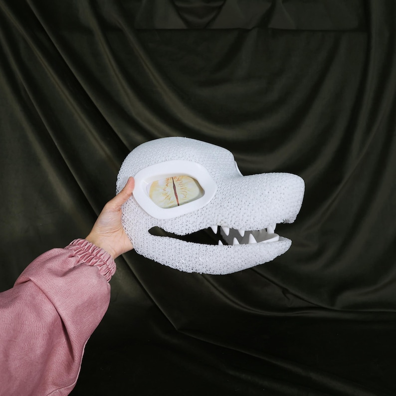 May include: A white, textured dragon head mask with large, oval eye openings. The mask features a detailed snout with sharp, white teeth. The eyes have a yellow and brown design. The mask is held by a person wearing a pink long-sleeved shirt.