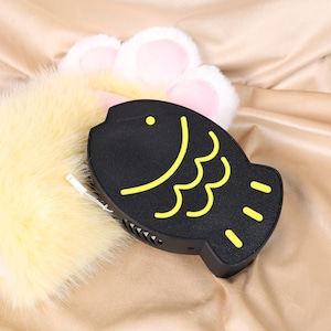 May include: A black, fish-shaped electronic device with yellow accents, resting on a plush, yellow surface. The device features a yellow fish outline and small vents. A fluffy, yellow paw is visible in the background.