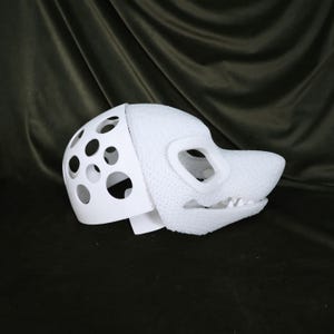 May include: A white, partially assembled fursuit head with a dog-like snout and open mouth. The head features a perforated section for ventilation and eye openings. The textured material contrasts with the smooth, solid white plastic.