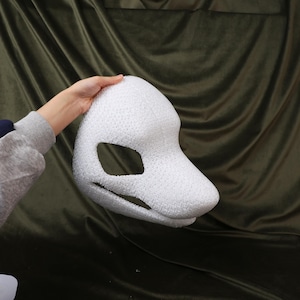 May include: A white, textured mask with a large eye opening and a snout-like shape. The mask is held by a person's hand against a dark green background. The mask appears to be made of a foam-like material.
