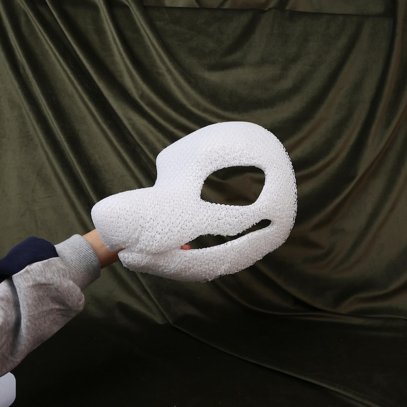 May include: A white, textured mask shaped like a stylized animal head. The mask has large eye openings and a long snout. The mask is held up by a person's arm, with a dark green background.