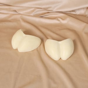 May include: Two off-white, heart-shaped objects are displayed on a beige, satin-like fabric. The objects have a smooth texture and are positioned separately, with one slightly angled towards the other.