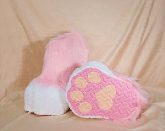 May include: Pink and white furry paw-shaped costume feet. The bottom of the paw is pink with a textured pattern and light orange paw pads. The top of the feet are pink with white tips.