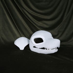 May include: A white, textured dragon head mask with large eye openings and a detailed jawline. A separate white piece, possibly a cheek or ear, sits to the left. The mask is likely for cosplay or costume use.