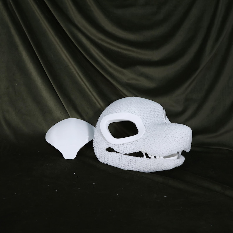 May include: A white, textured dragon head mask with large eye openings and a detailed jawline. A separate white piece, possibly a cheek or ear, sits to the left. The mask is likely for cosplay or costume use.