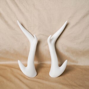 May include: A pair of white, smooth, curved mannequin hand displays. Each piece features an elongated, tapered finger pointing upward and a shorter, curved thumb-like projection at the base, designed for showcasing jewelry or accessories.