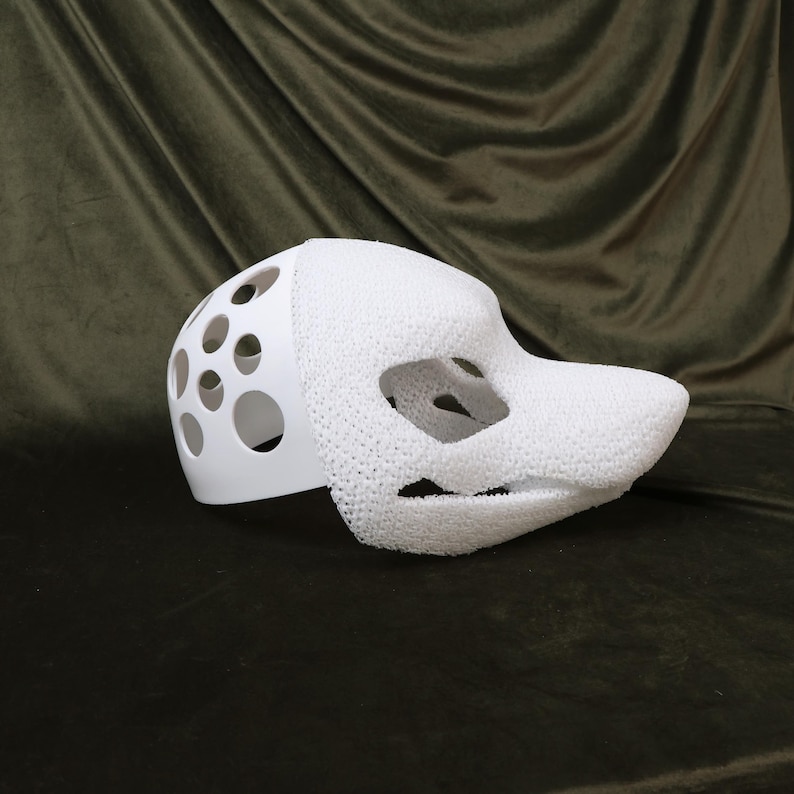 May include: A white, partially assembled fursuit head base. The base features a smooth, solid white section with circular cutouts and a textured, foam-like snout and face. The design includes eye and mouth openings.