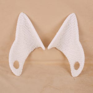 Puede incluir: Dos accesorios blancos, texturizados, con forma de orejas de gato. Cada oreja tiene una base redondeada con un pequeño agujero y termina en punta. Las orejas están hechas de un material tejido, sobre un fondo neutro.