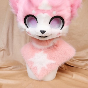 Pode incluir: Uma cabeça de fursuit rosa e branca com olhos grandes e expressivos e um rosto sorridente. A cabeça apresenta um desenho em forma de estrela no pescoço e foi projetada para ser usada. As orelhas são rosa e os olhos são grandes e redondos.