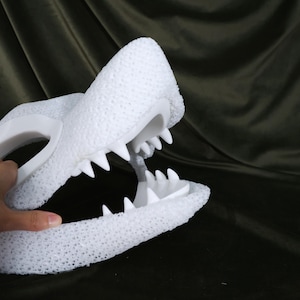 May include: A white, textured, open-mouthed alligator or crocodile head with sharp, pointed teeth. The head is held by a hand, and the background is a dark, textured fabric.