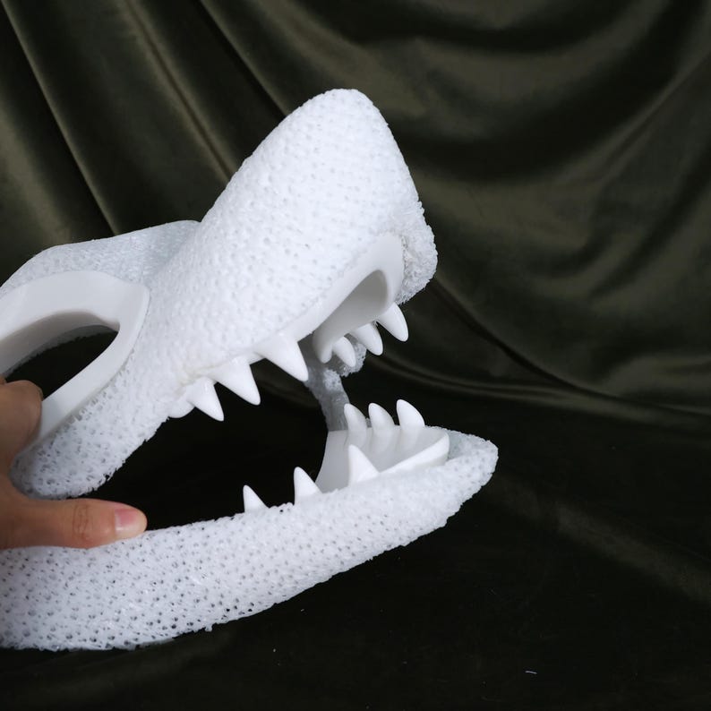 May include: A white, textured, open-mouthed alligator or crocodile head with sharp, pointed teeth. The head is held by a hand, and the background is a dark, textured fabric.