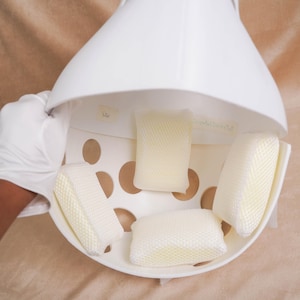 May include: A white, dome-shaped hair dryer hood attachment featuring a series of circular ventilation holes. The interior is lined with three rectangular, pale yellow, mesh-covered foam cushions positioned to provide comfort during use.