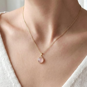 May include: A gold-coloured necklace with a small, round, pink gemstone pendant. The pendant hangs from a fine chain. The necklace is shown against a neutral background, emphasising the jewellery's simple elegance.