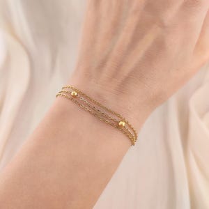 Può includere: Un delicato braccialetto color oro caratterizzato da tre catenine sottili sovrapposte. Il design include due piccole perline sferiche dorate e lisce distanziate lungo le catene, creando un aspetto a strati con una finitura metallica lucida e maglie fini.