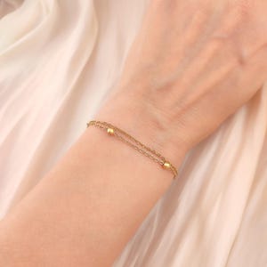 Può includere: Un delicato bracciale a doppia catena color oro caratterizzato da due piccole perle sferiche e lisce distanziate lungo le maglie. Il bracciale è realizzato con sottili maglie di catena metallica che avvolgono il polso.